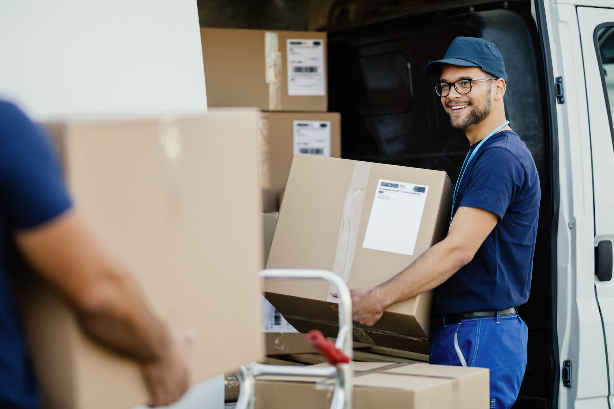 Joven trabajador manual feliz que lleva cajas de carton en la furgoneta de reparto mientras se comunica con sus colegas 2